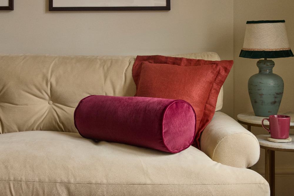 A home interior scene showing a cream tufted sofa with two decorative pillows: a cylindrical magenta pillow in front and a rectangular rust-orange pillow behind it. A side table with a blue-green lamp and a pink mug sits to the right.