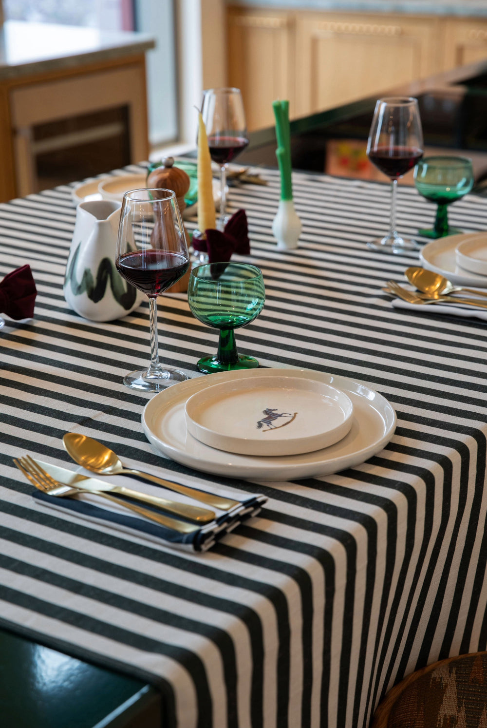 A black and white striped tablecloth draped over a dining table, styled with white plates, gold cutlery, wine glasses, and green glass accents in a warm indoor setting.