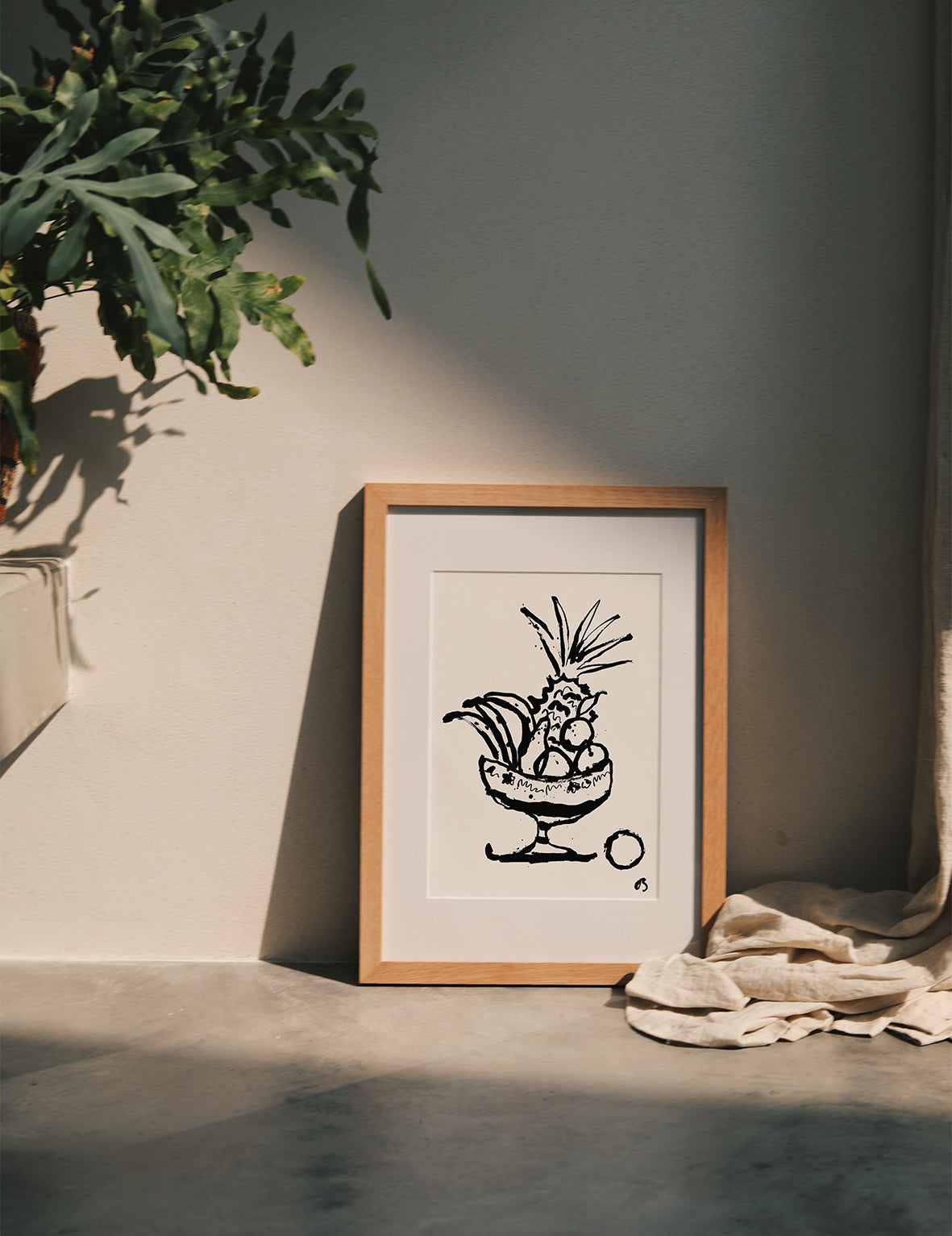 Framed fruit bowl artwork on a neutral background with a plant and fabric.