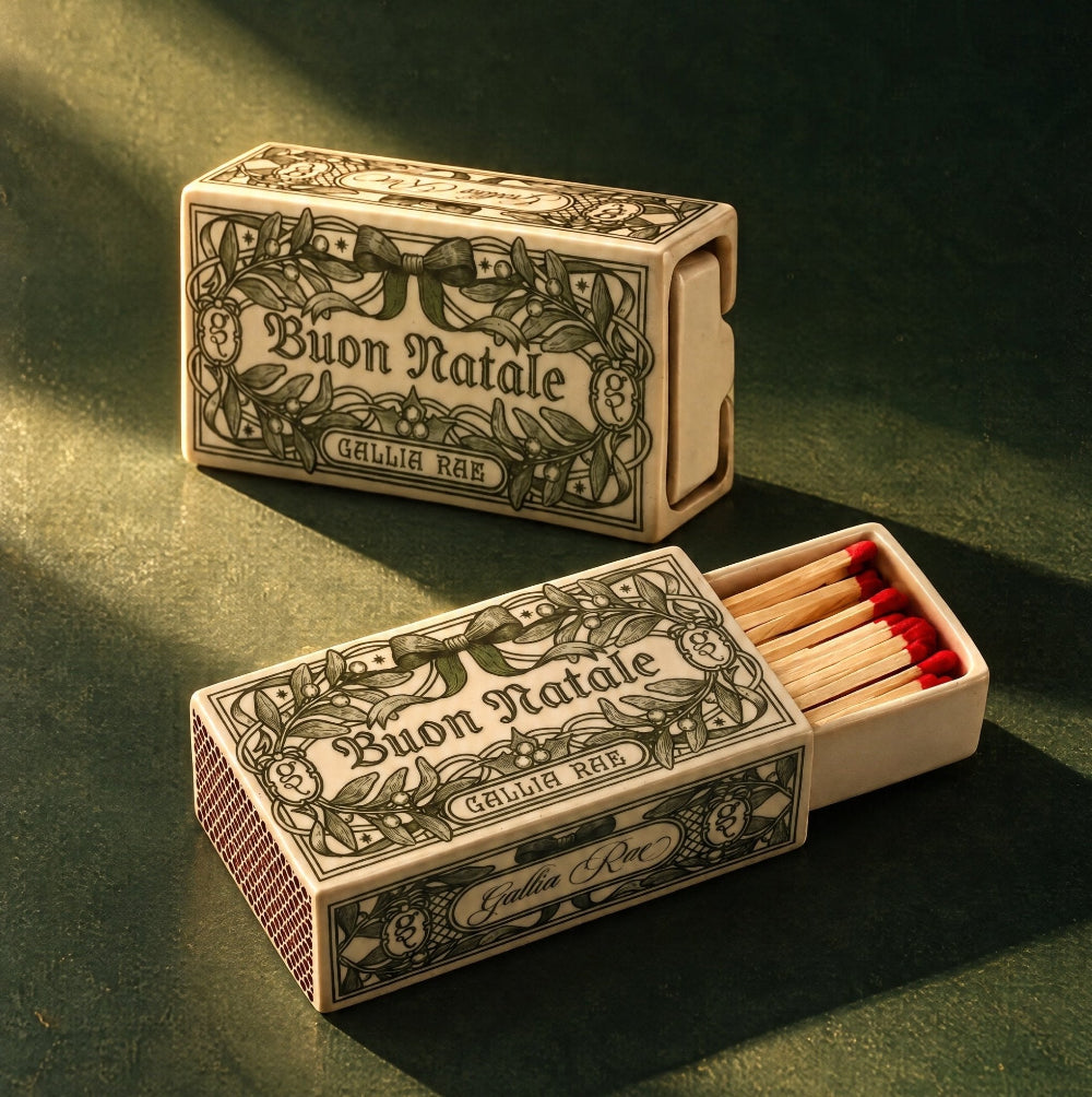 Two Gallia Rae Buon Natale ceramic matchboxes on a dark green background, one open with matches inside