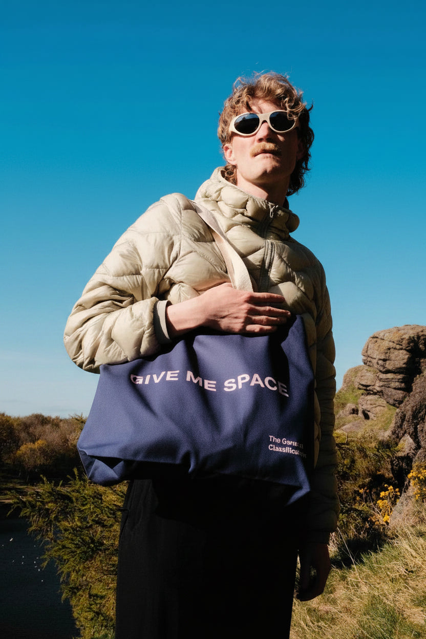 A person outdoors wearing a beige puffer jacket and sunglasses, carrying a navy tote bag with the text 'GIVE ME SPACE' and 'The Gave Classifications' on it. The background shows a blue sky and rocky landscape.