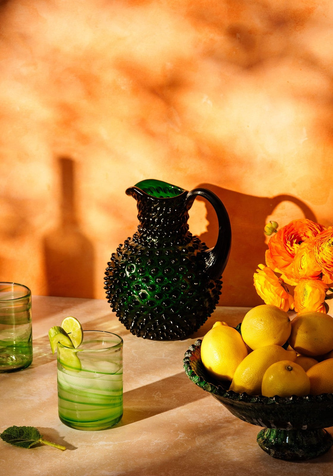 Green glass pitcher and glass with lemon slices on a wooden surface with a warm-toned background.