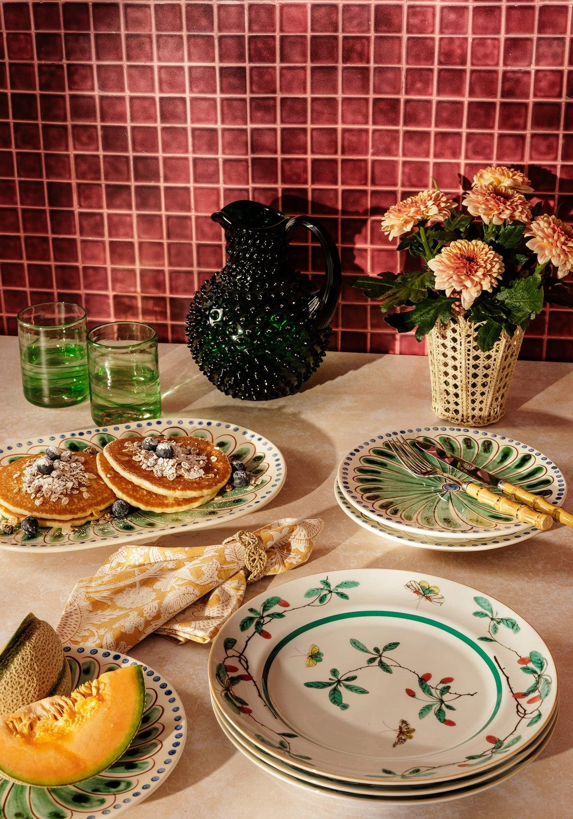 Table setting with pancakes, fruit, and decorative plates against a tiled wall.