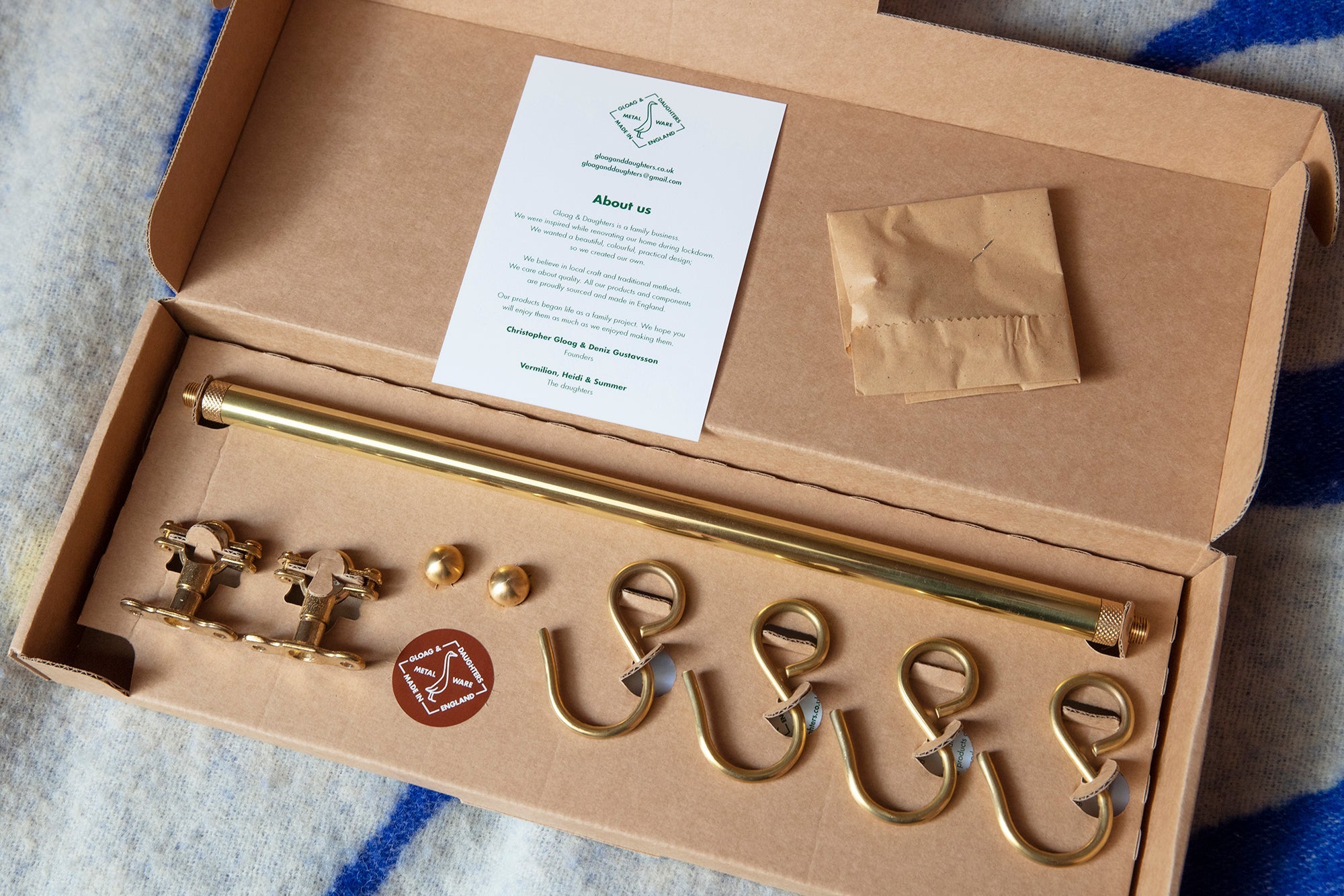 A brass kitchen rod set with five matching hooks, two small gold balls, and a white instruction card inside an open brown cardboard box, placed on a blue and white patterned blanket.