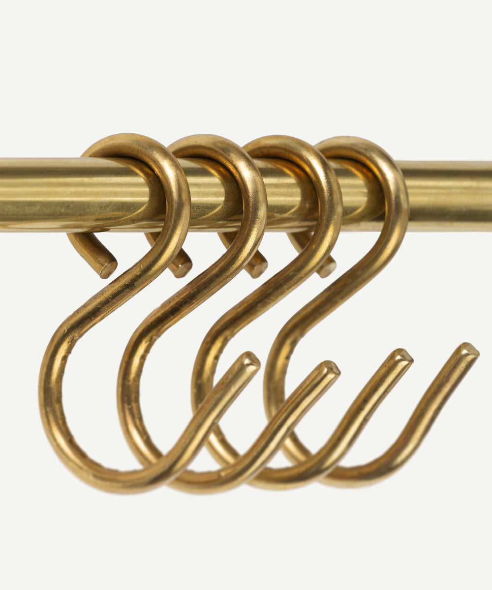 Four brass S-hooks hanging from a matching brass rod against a plain off-white background.