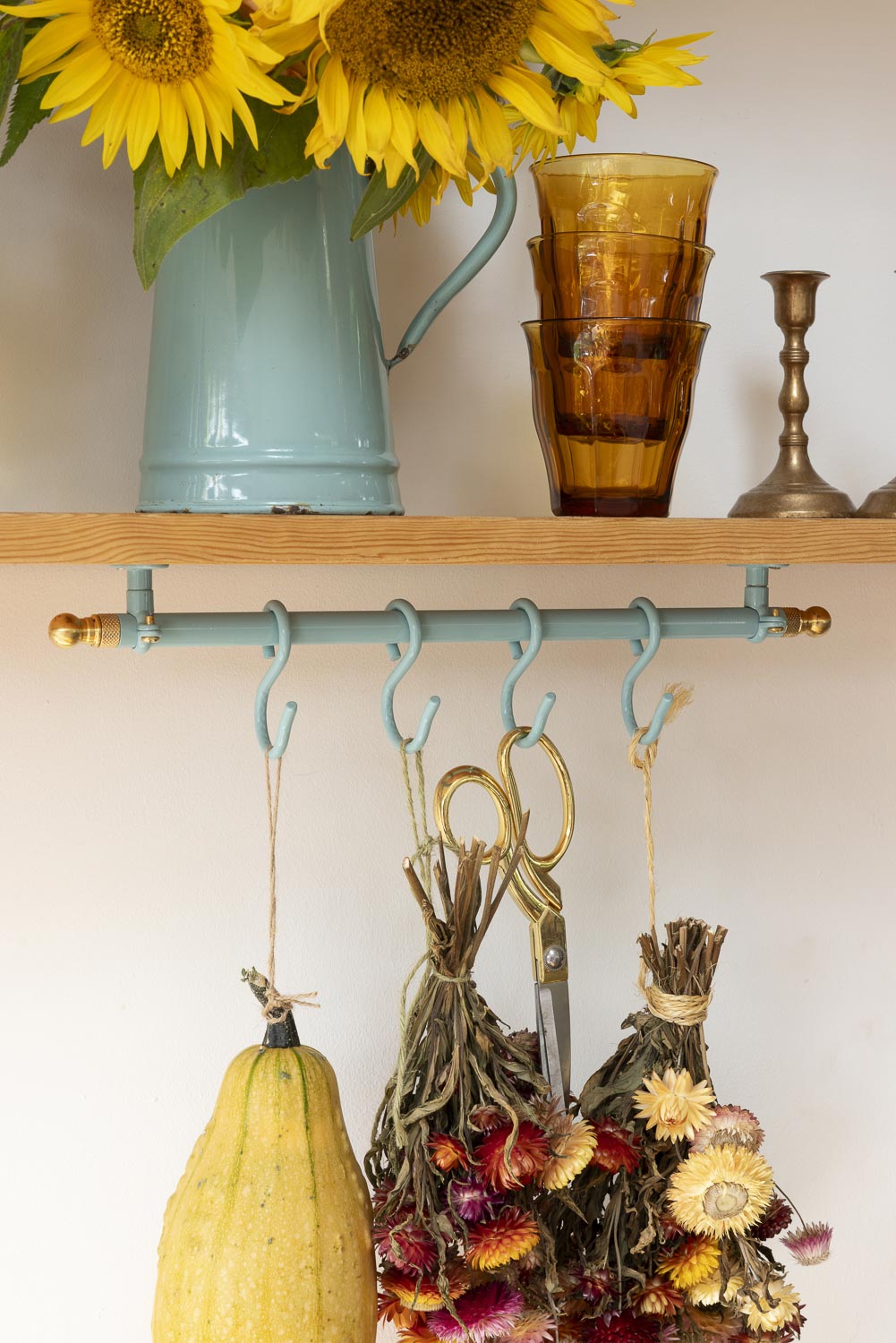 A rustic teal wall-mounted hook rack with brass-toned hardware, shown holding dried flower bundles, scissors, and a small yellow squash. Above it sits a wooden shelf with a light blue pitcher of sunflowers and amber glass cups.