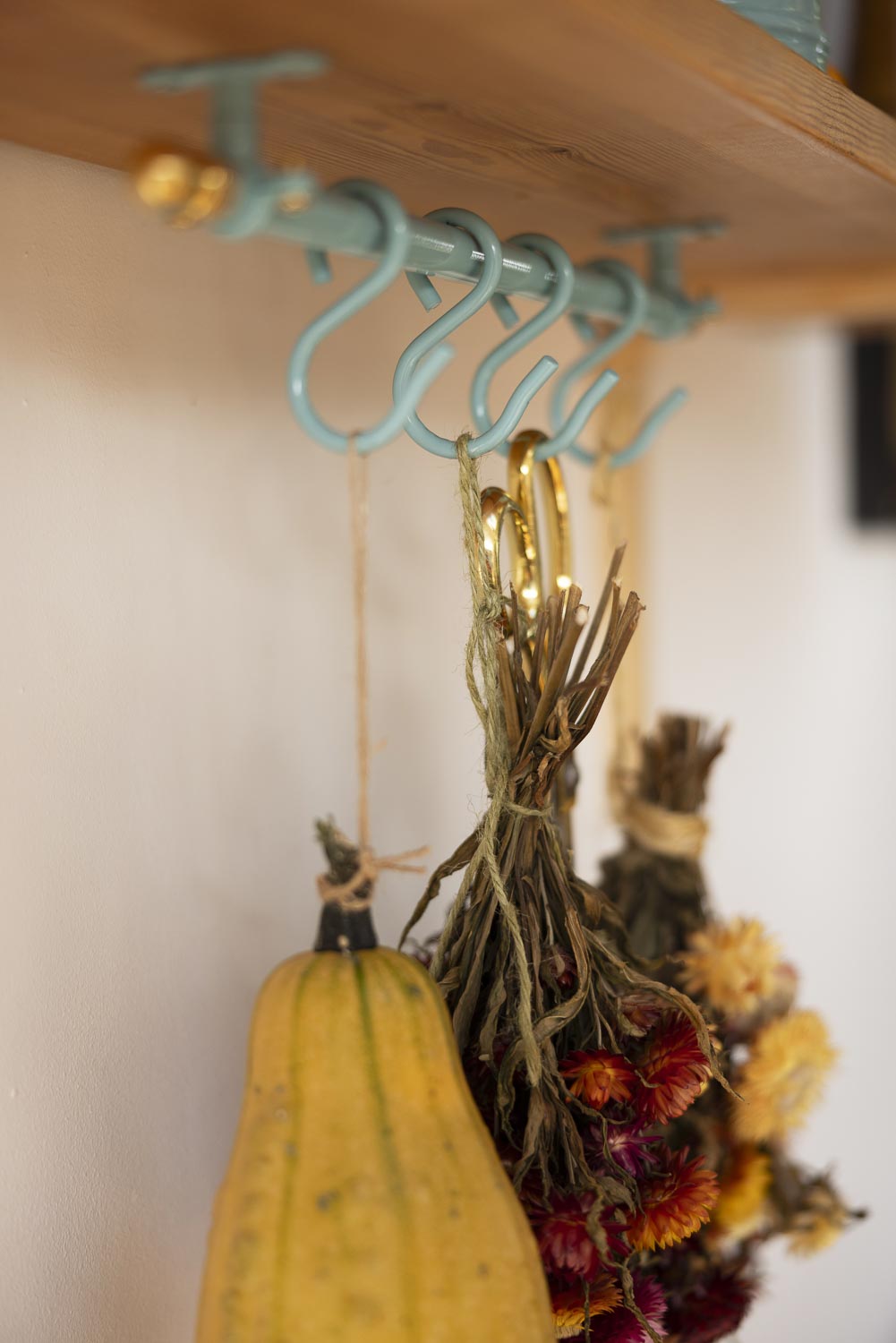 A close-up of a teal metal hanging rack mounted on a wooden shelf, with dried flower bundles and a yellow-orange gourd hanging from the hooks in a warm indoor setting.