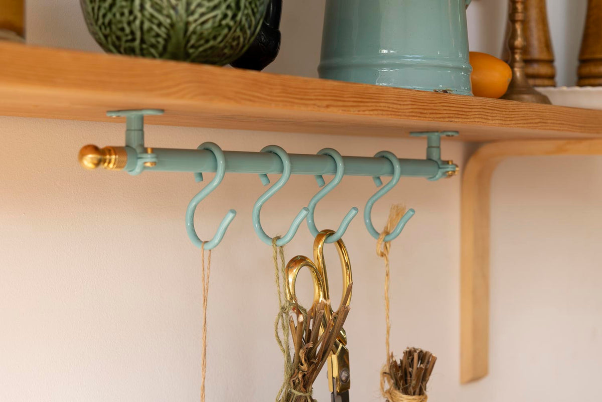 A teal metal wall-mounted hanging rack with five hooks, shown mounted on a wooden shelf with decorative items and hanging accessories below.