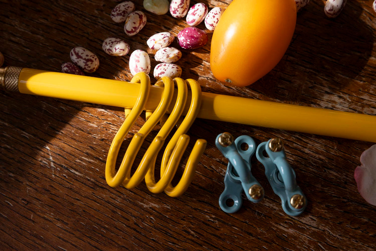A yellow metal S-hook rack with two matching blue mounting brackets placed on a rustic wooden surface, alongside decorative beans and a yellow fruit.