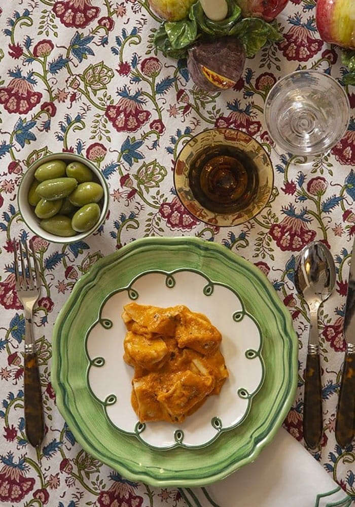Dish of food on a patterned tablecloth with a bowl of pickles and a glass of water.