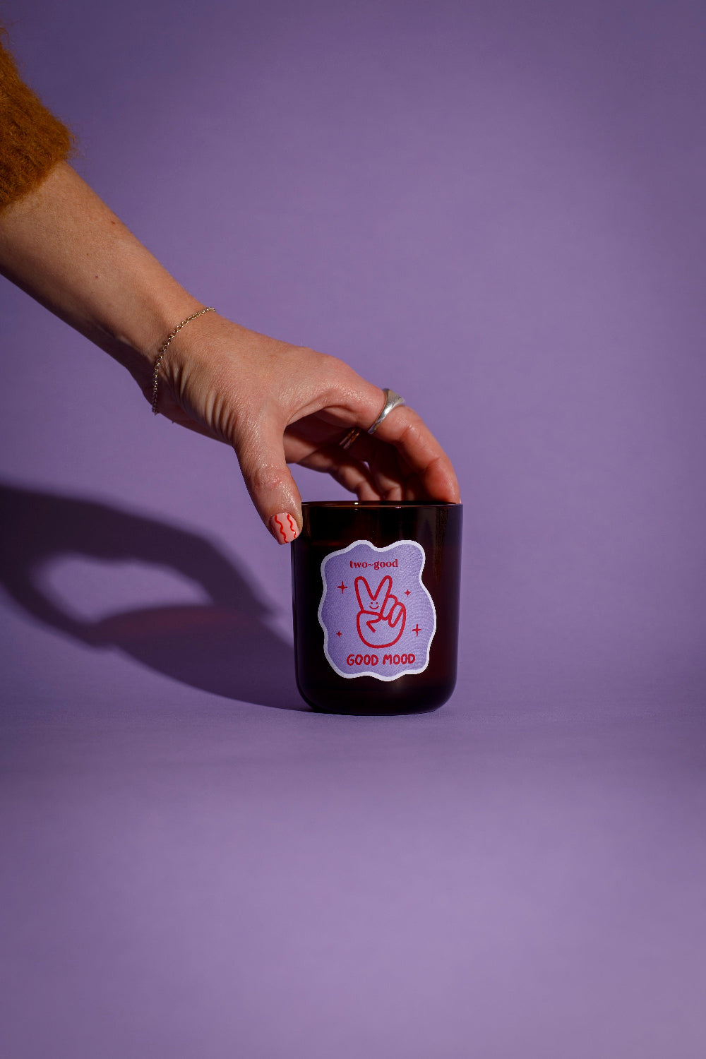 Hand holding a candle jar with a pink and white label called 'Good Mood' against a purple background
