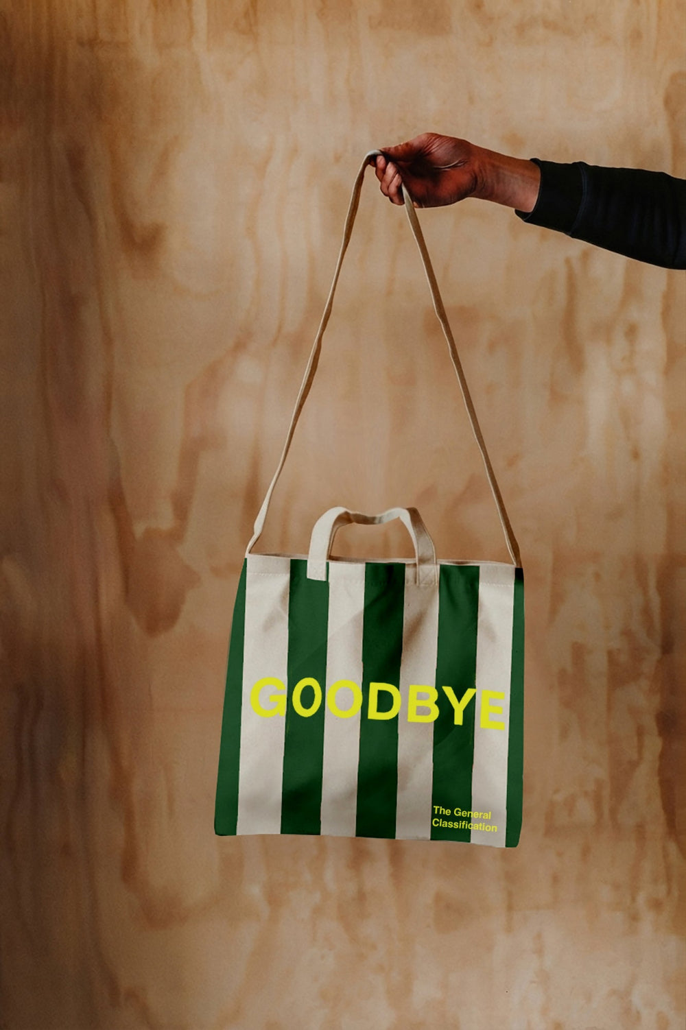 A green and white striped canvas tote bag with yellow 'GOODBYE' text, held by a hand against a warm wood background.