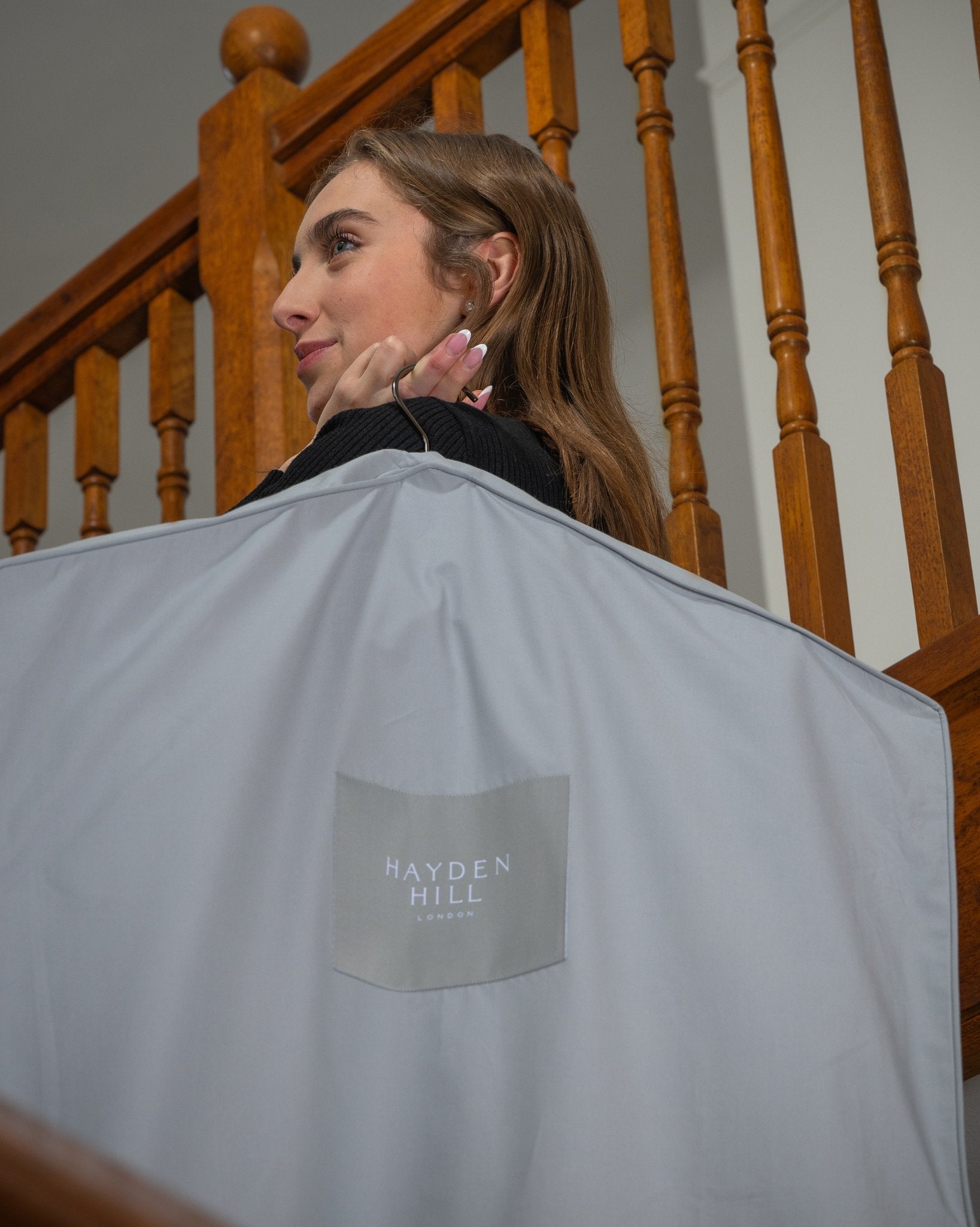 Person holding a grey garment bag with 'Hayden Hill' branding in front of wooden stairs.