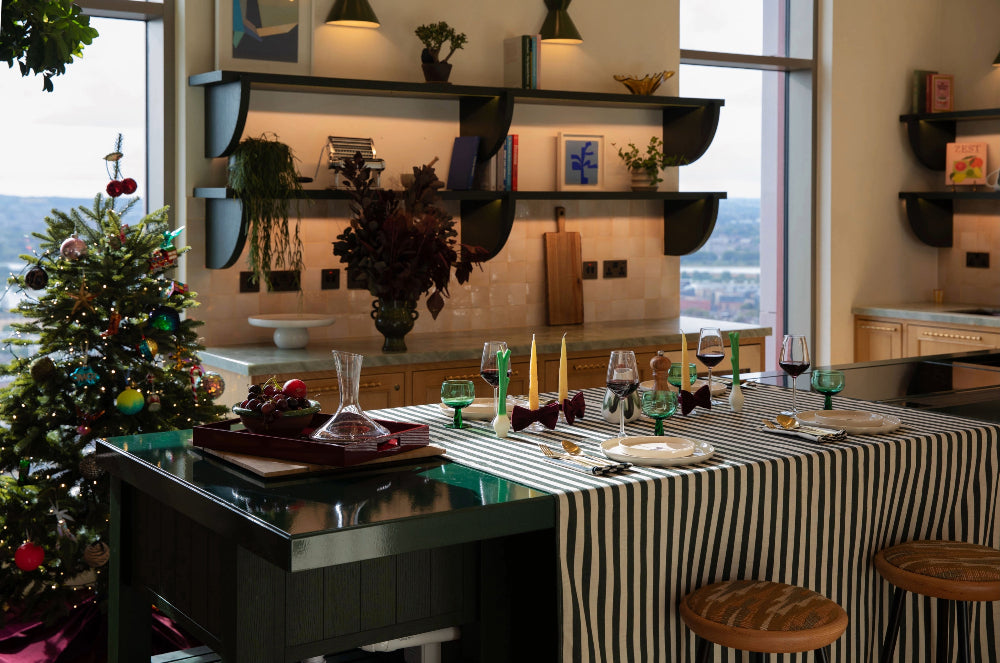 A styled dining table in a modern kitchen setting, covered with a black and white striped tablecloth and set with wine glasses, candles, and a carafe. A decorated Christmas tree stands to the left, and large windows in the background show an outdoor view.