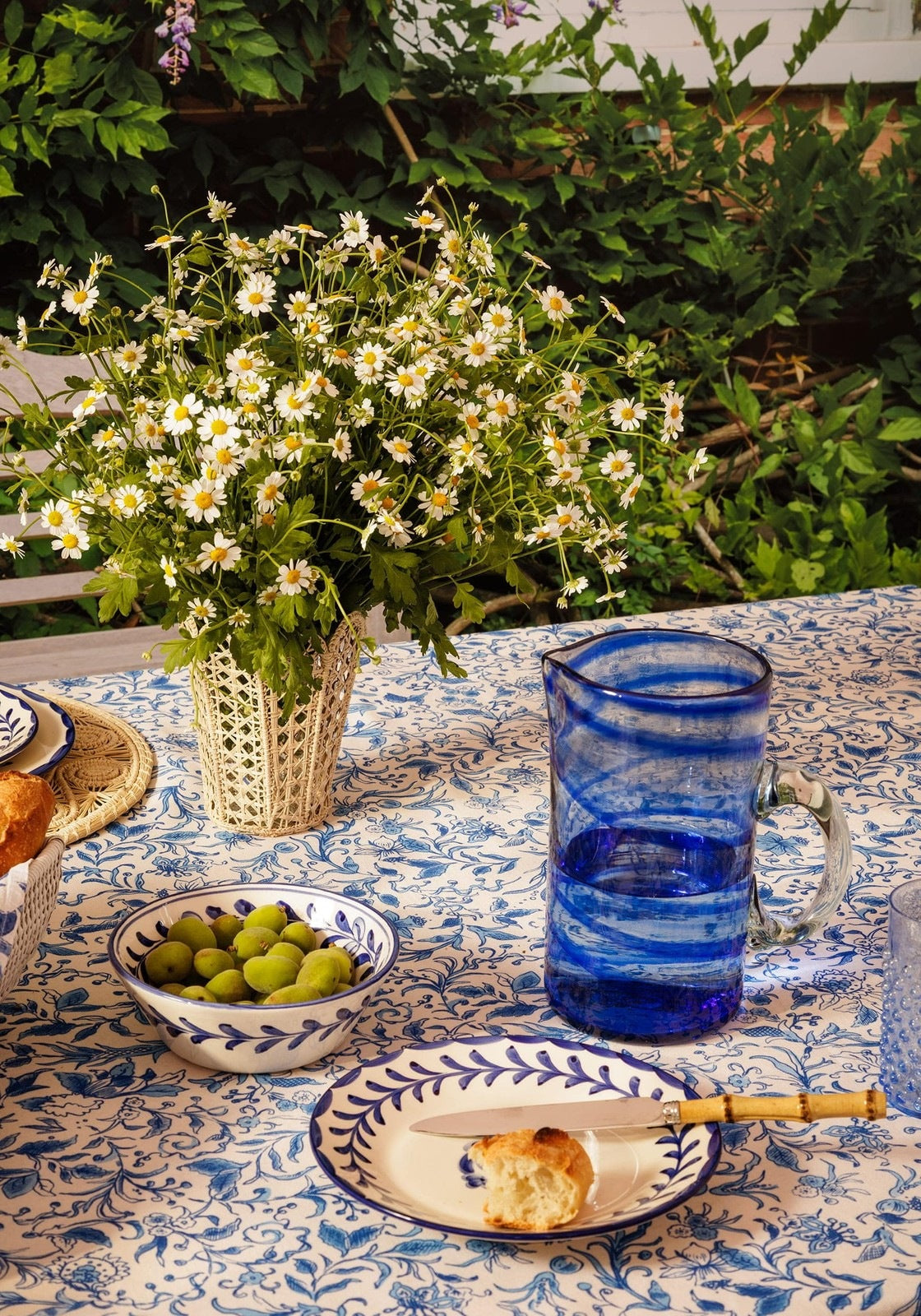 Outdoor table setting with a blue handblown glass pitcher, greenery, and food on a patterned tablecloth.