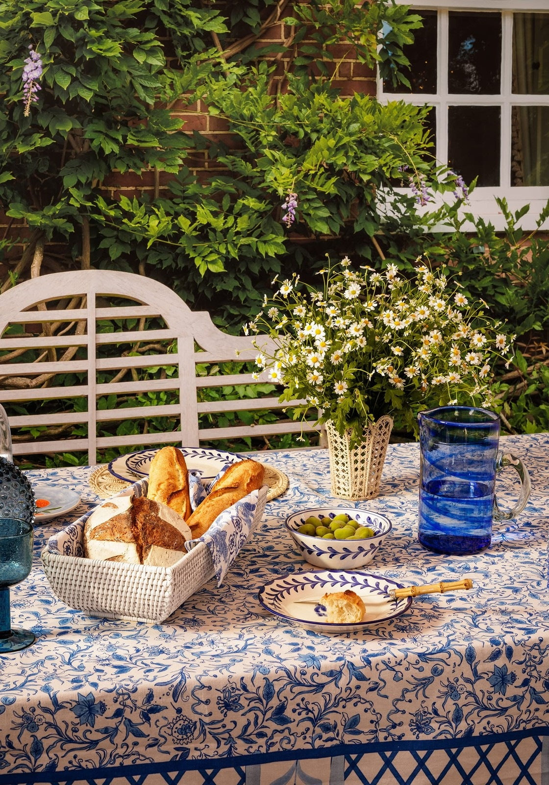 Outdoor dining table with bread basket, plates, and glasses in a garden setting.