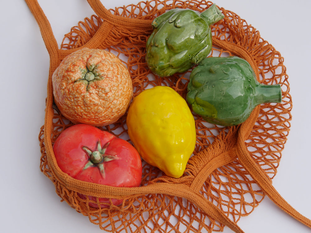 Colourful vegetables including a tomato, yellow pepper, and green artichokes on an orange woven mesh bag.