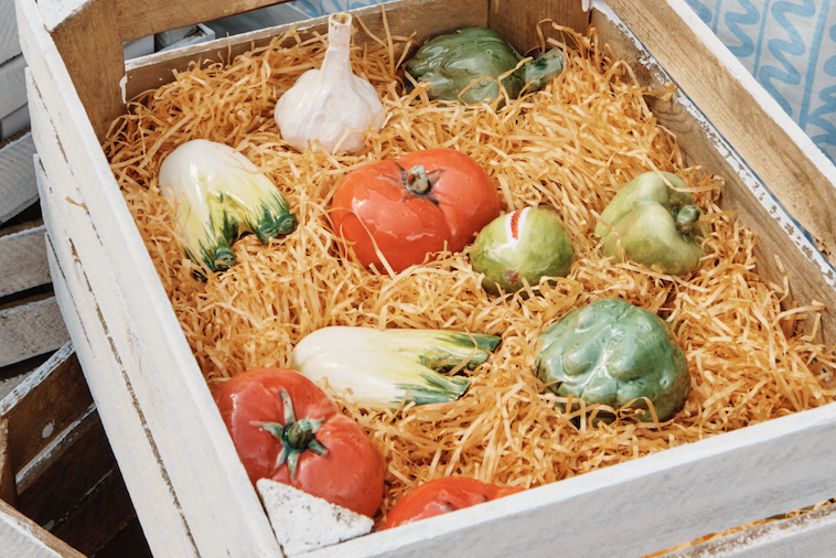 Box filled with straw and ceramic vegetables including tomatoes and green peppers.