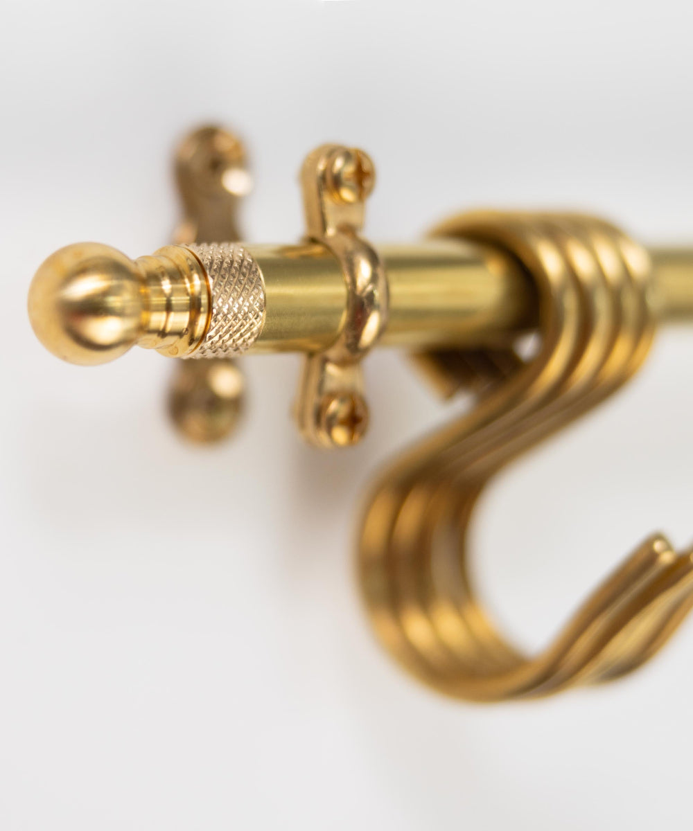 Close-up of a polished brass curtain rod with a decorative crossbar and a large spiral hook, shown against a plain white background.