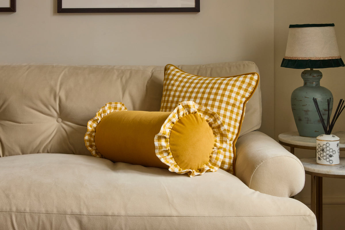 A cozy living room scene featuring two decorative throw pillows on a beige sofa: a round yellow velvet lumbar pillow with ruffled trim and a square yellow-and-white gingham pillow, next to a side table with a lamp.