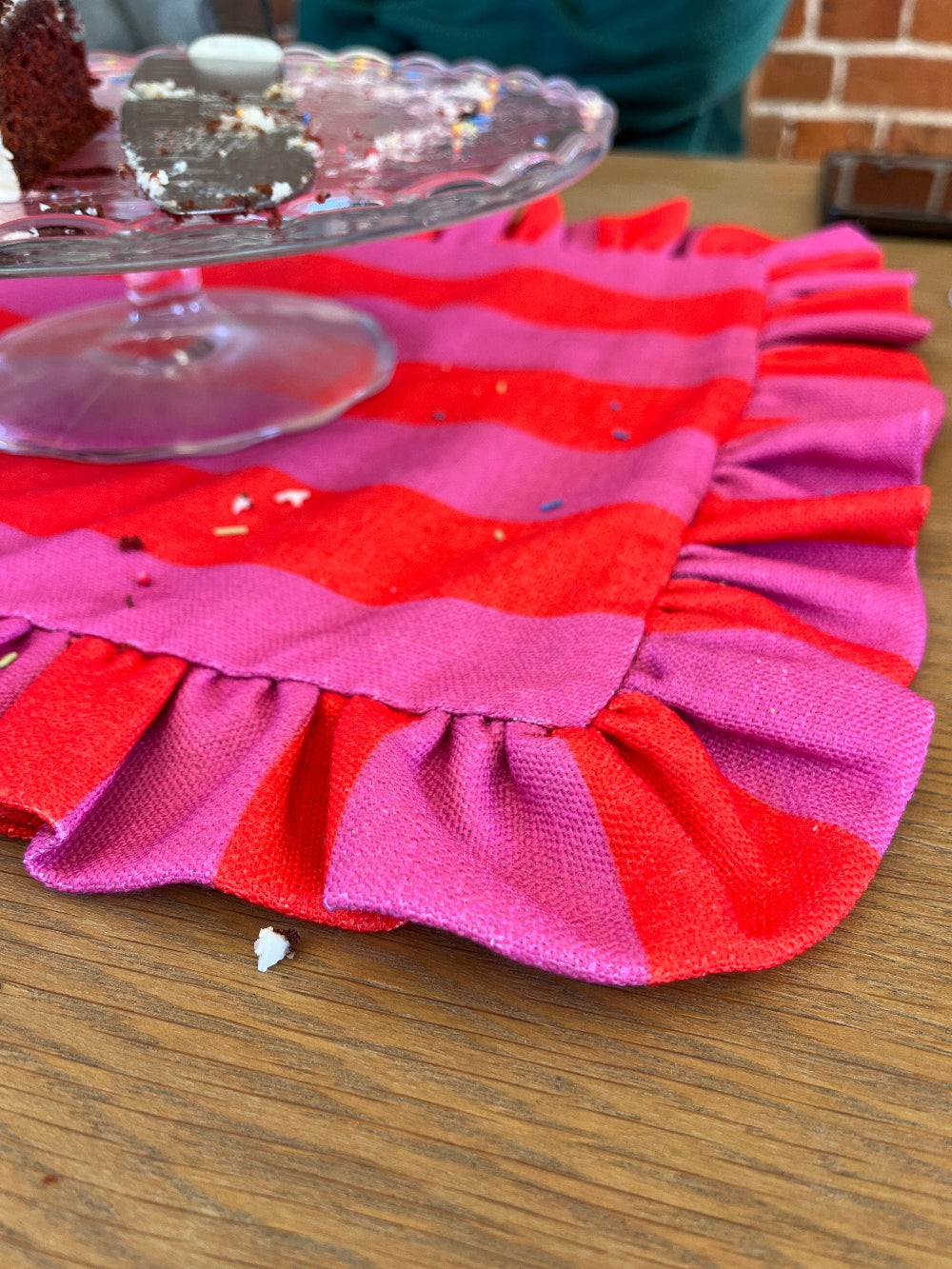 A close-up of a pink and red striped fabric table runner with ruffled edges on a wooden table, with a glass cake stand partially visible in the background.