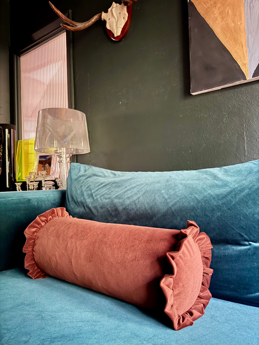 A dusty rose velvet cylindrical lumbar pillow with ruffled trim resting on a teal velvet sofa in a styled living room setting.