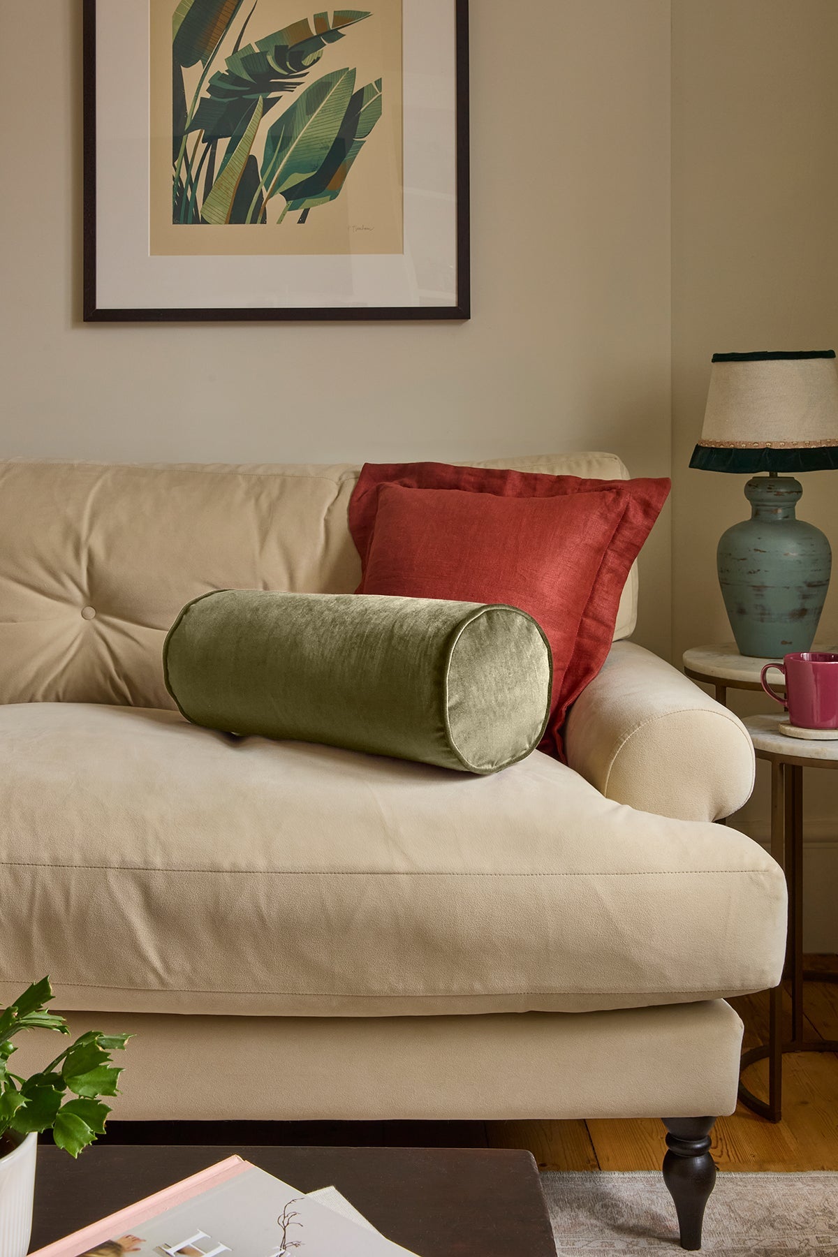 A styled living room scene featuring a cream-colored sofa with two decorative pillows: a cylindrical sage green velvet pillow in front and a square red throw pillow behind it. A framed botanical print hangs on the beige wall above, and a blue-green table lamp sits on a side table to the right.