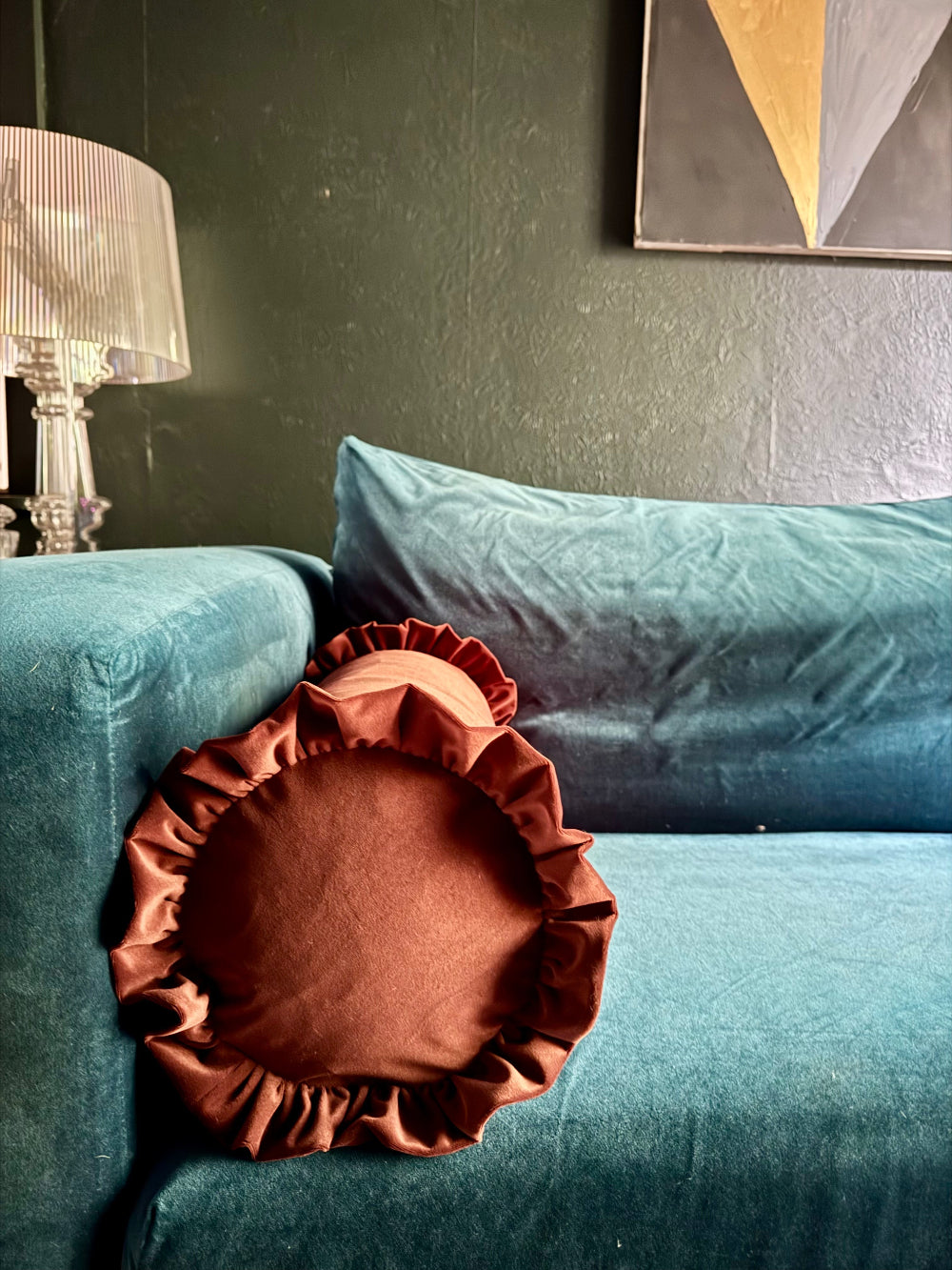 A rust-brown ruffled accent pillow resting on a teal velvet sofa in a styled living room setting.