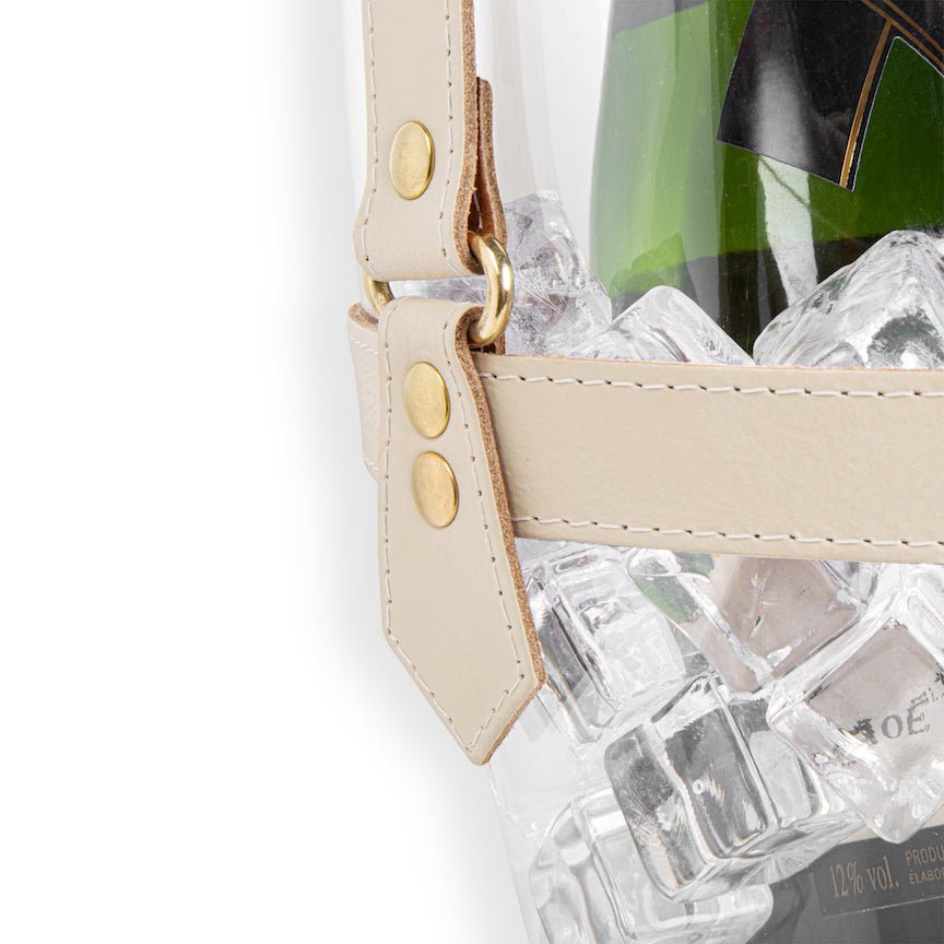 Clear champagne bucket with beige strap containing a bottle of champagne and ice cubes on a white background