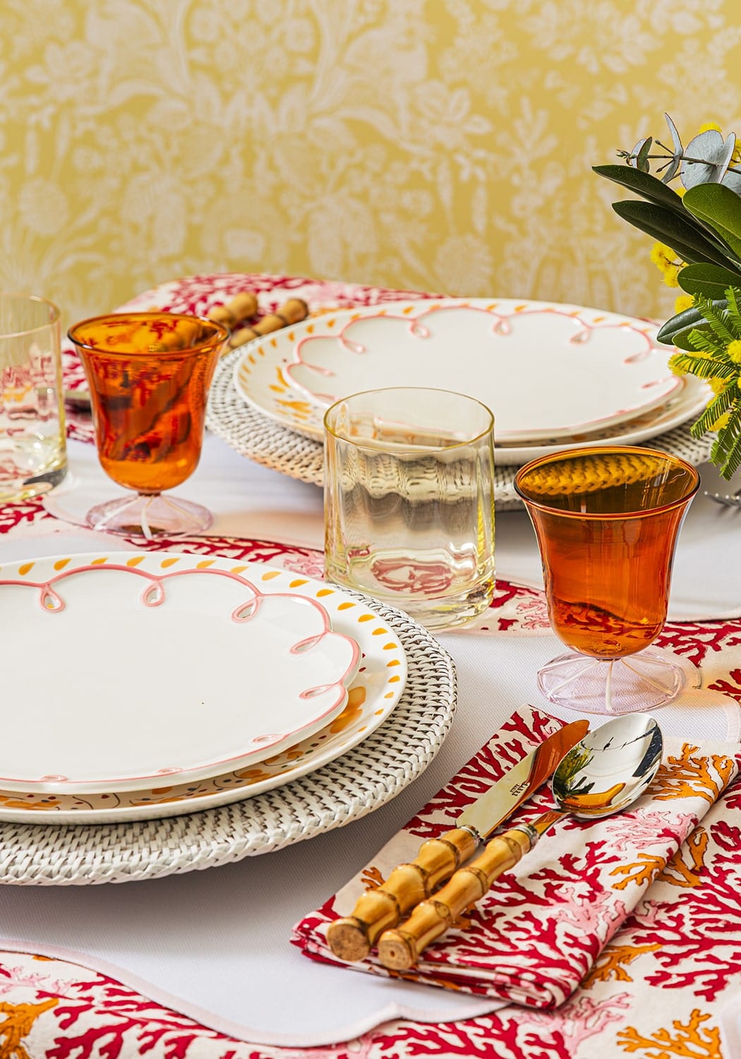 Dining table setting with colorful plates, glasses, and cutlery on a patterned tablecloth.