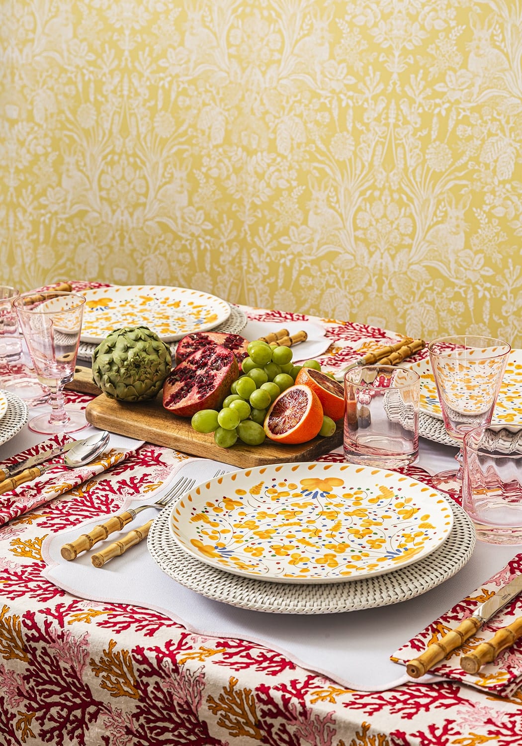 Dining table setting with floral plates, fruits, and cutlery on a patterned tablecloth.