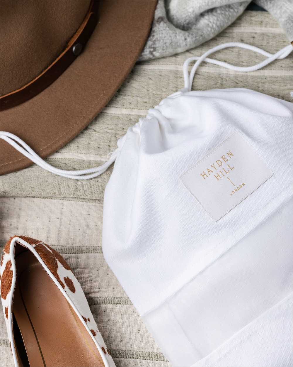A white drawstring shoe bag with a square beige Hayden Hill label, styled in a flat lay with a brown hat, gray knit, and a brown-and-white patterned shoe on a light wood surface.