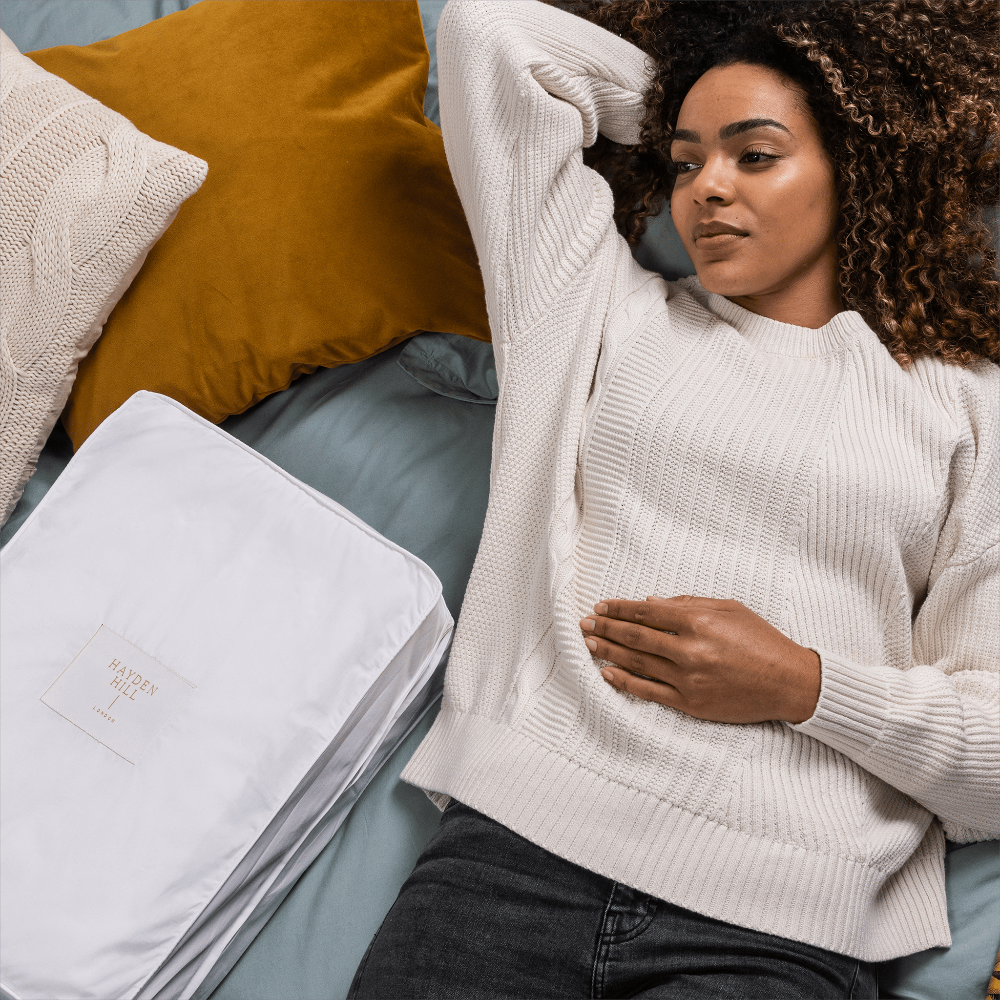 Woman in a white sweater sitting on a bed with pillows and a white storage bag.