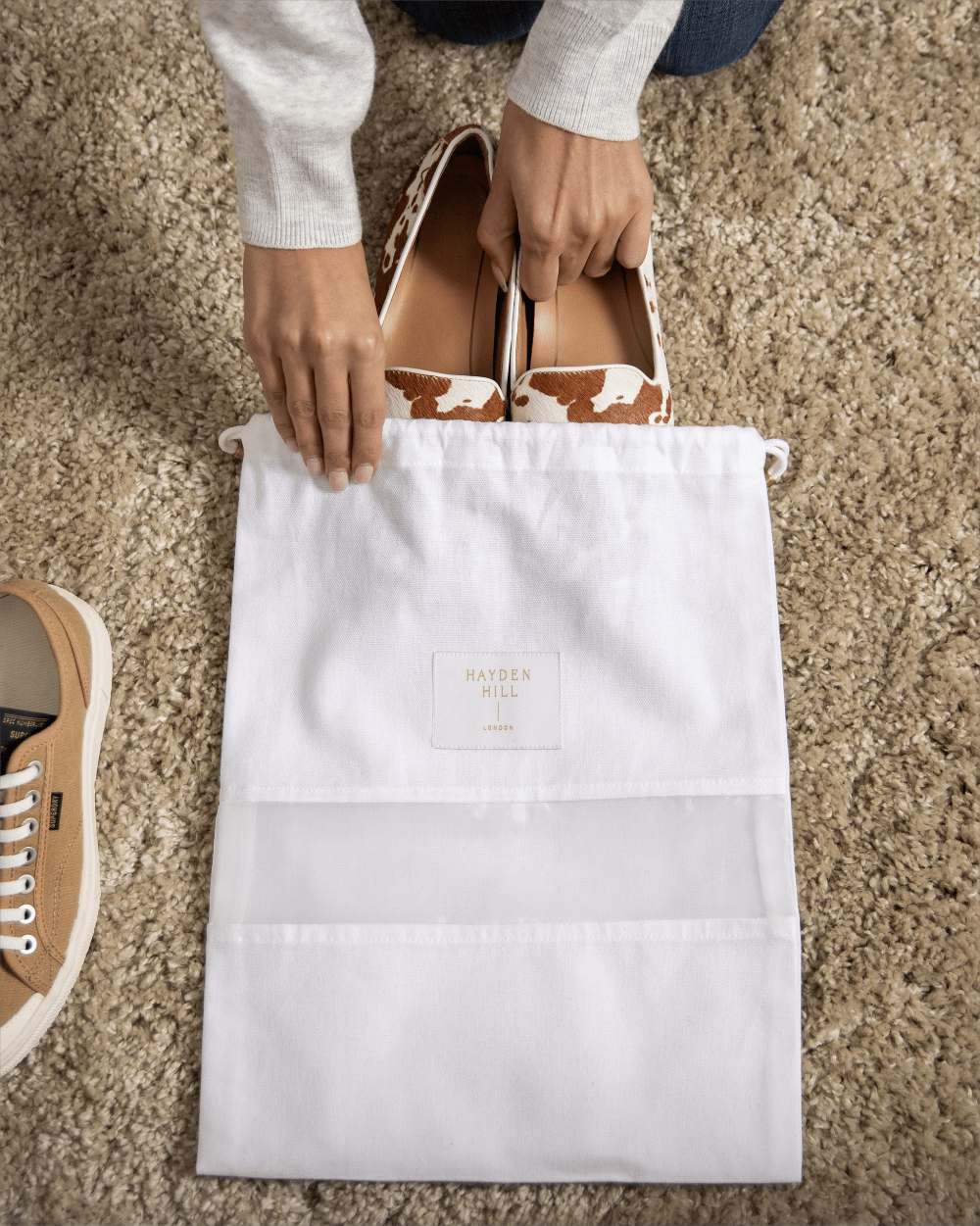 A white drawstring shoe bag with a small 'HAYDEN HILL' label in the centre, being held open by hands with brown-and-white patterned shoes partially inside. The scene is photographed from above on a beige carpet, with part of a tan sneaker visible at the left edge.