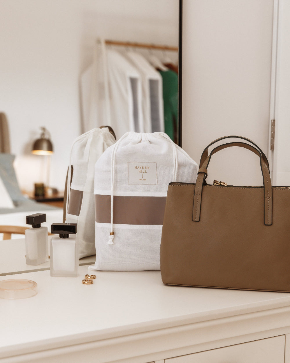 A styled collection of travel accessories on a white vanity table, featuring a brown leather tote bag, a white drawstring backpack with a brown stripe, and a white drawstring pouch, alongside perfume bottles and rings.