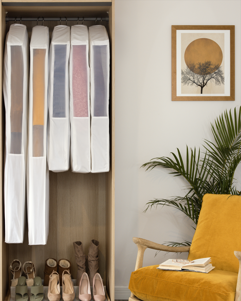Closet with hanging clothes and shoes, yellow chair with books, and framed artwork on the wall.