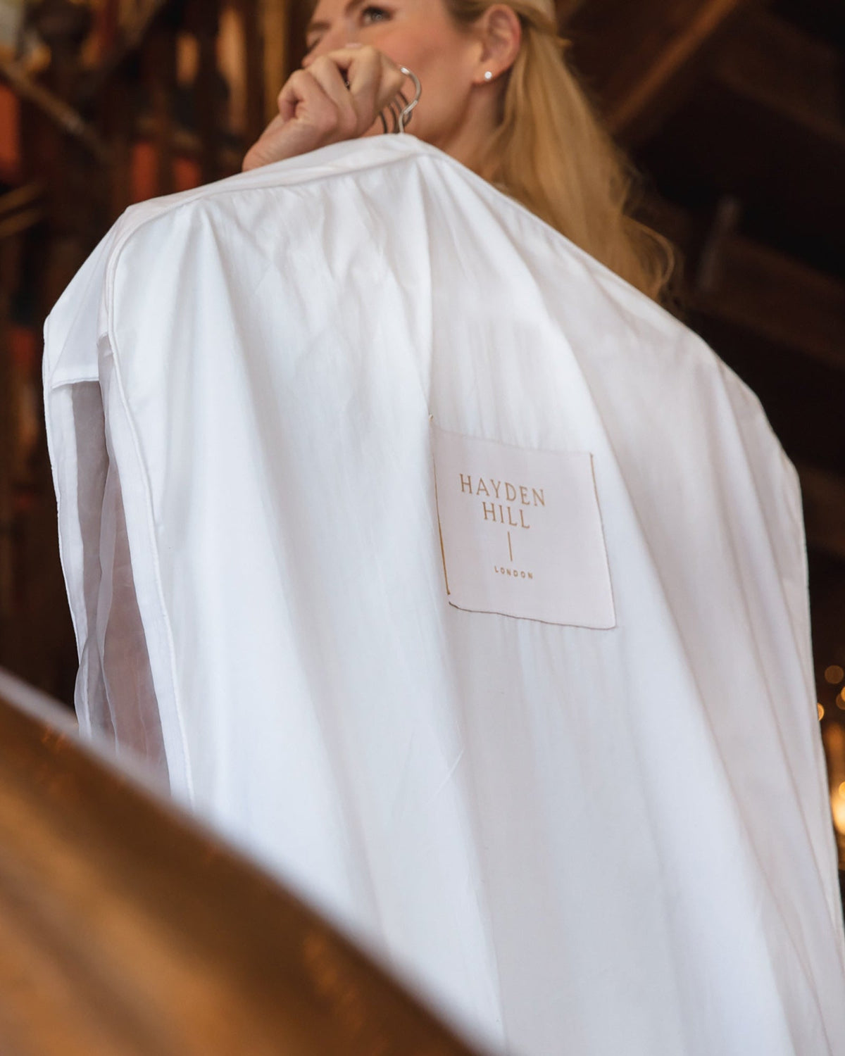 White hanging garment bag with 'Hayden Hill' label held by a person in a blurred indoor setting