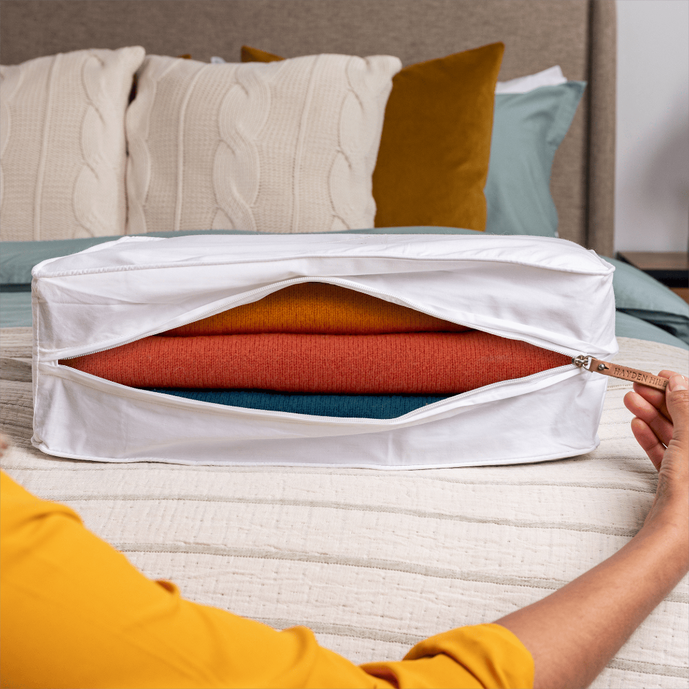 Person using a white storage bag to organize folded clothes on a bed.