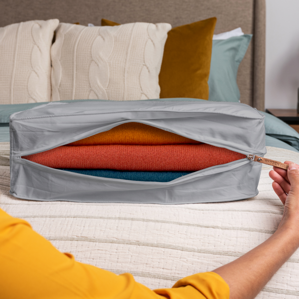 Person opening a grey storage bag containing colourful folded clothes on a bed.