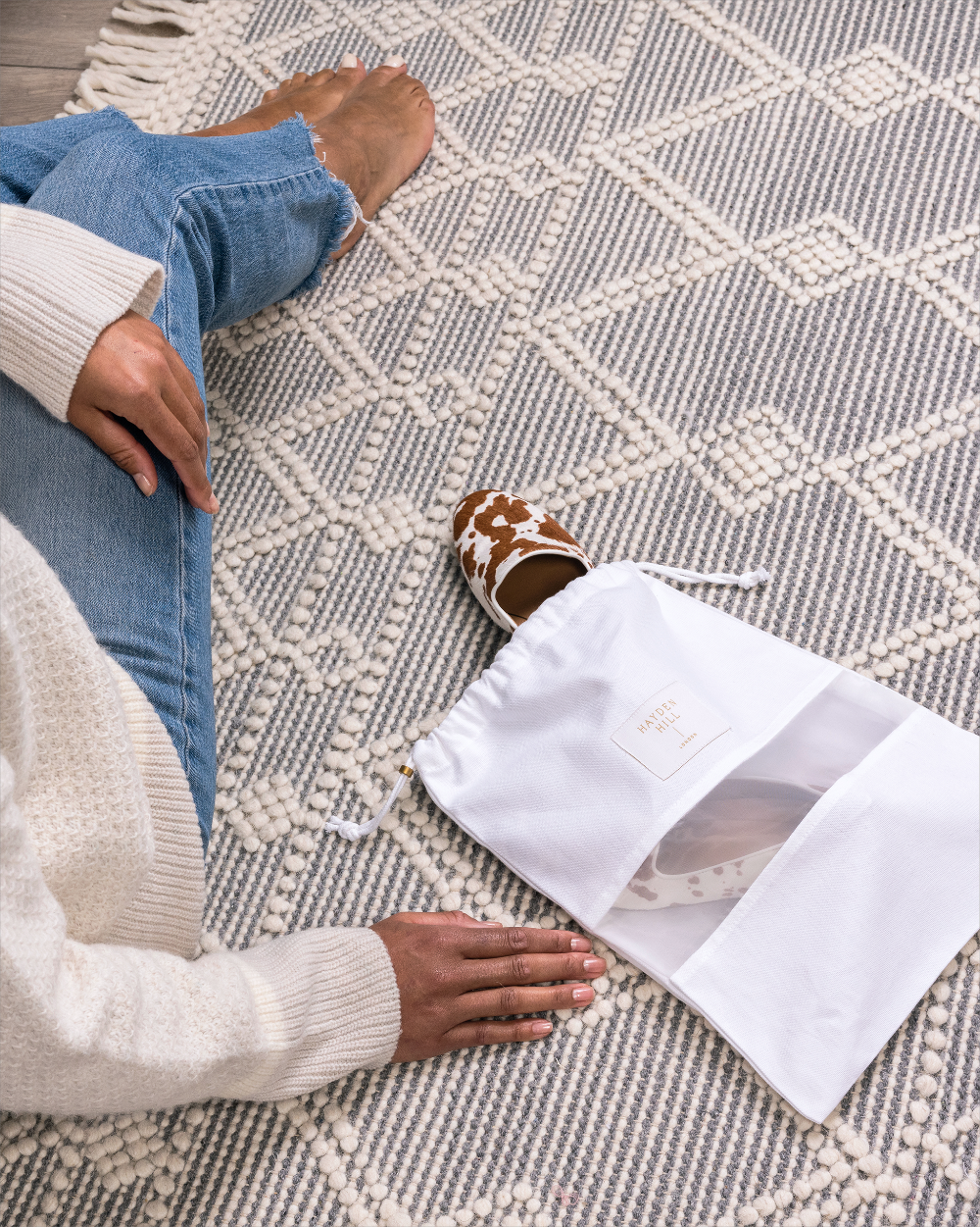 A person sitting on a patterned rug wearing brown and white cow-print slip-on shoes next to a white drawstring shoe bag with a clear window.