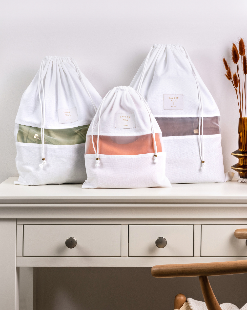 Three white drawstring storage bags with semi-transparent mesh bands are arranged on a white table. The bags have small rectangular labels and are shown in different colored interior compartments: olive green, peach, and dusty rose.