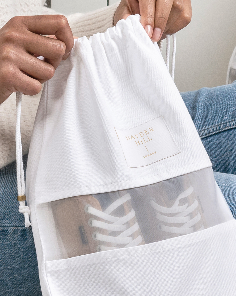 A white drawstring shoe bag with a clear front window showing white sneakers inside, held by hands over a person's lap. The bag has a square patch reading 'HAYDEN HILL' and is worn over blue jeans.