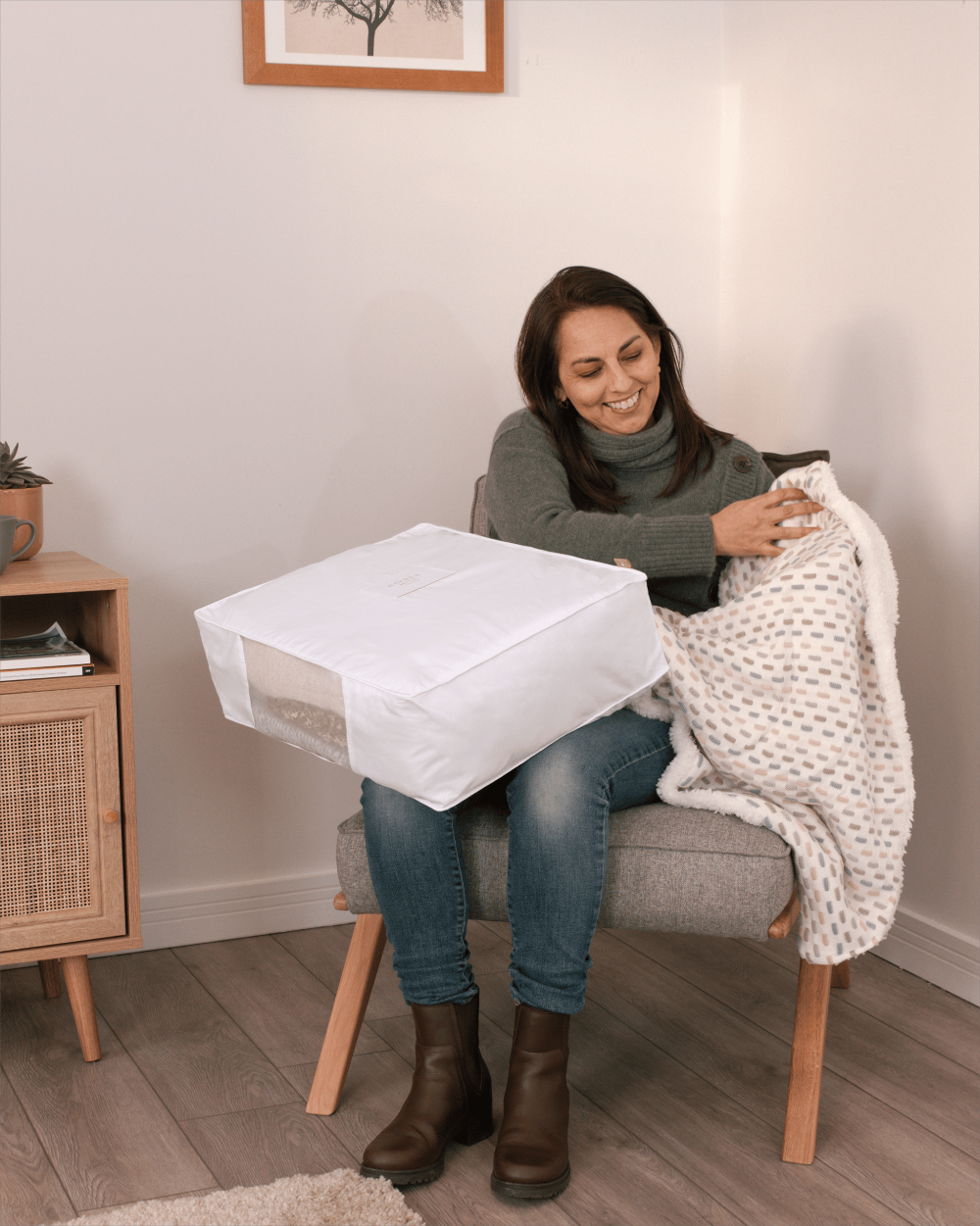 Woman sitting on a chair holding a white storage bag and a folded blanket in a room with a wooden cabinet and framed picture on the wall.