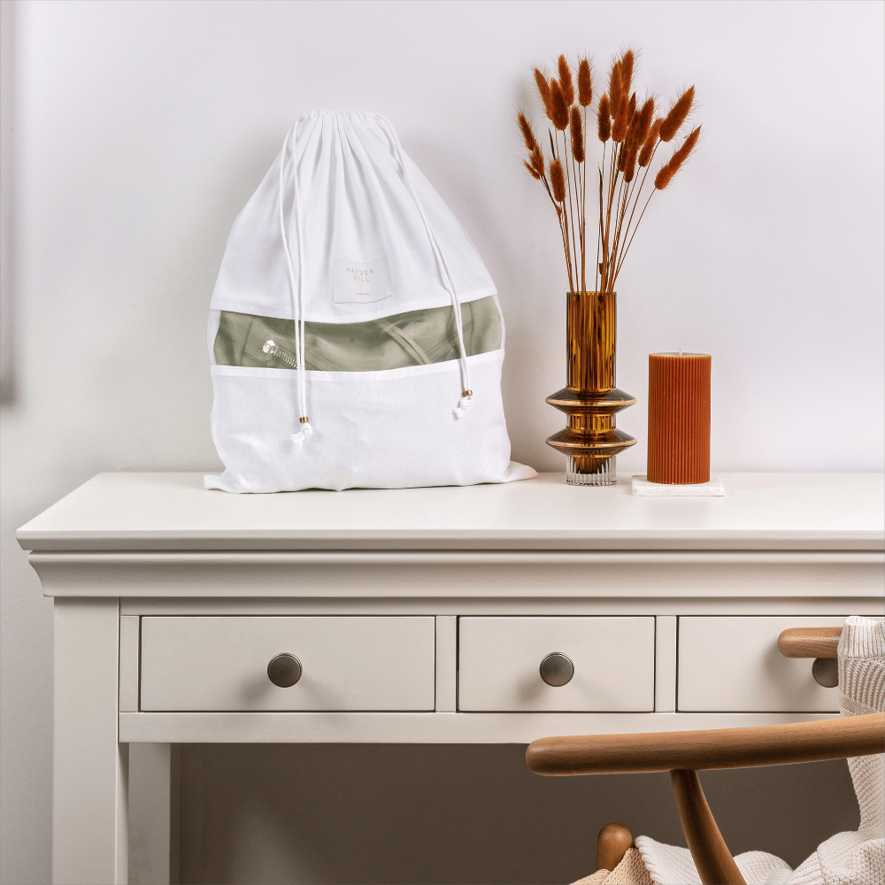 A white drawstring bag with a semi-transparent olive green band sits on a white wooden desk next to a decorative vase with dried grasses and a small orange candle.
