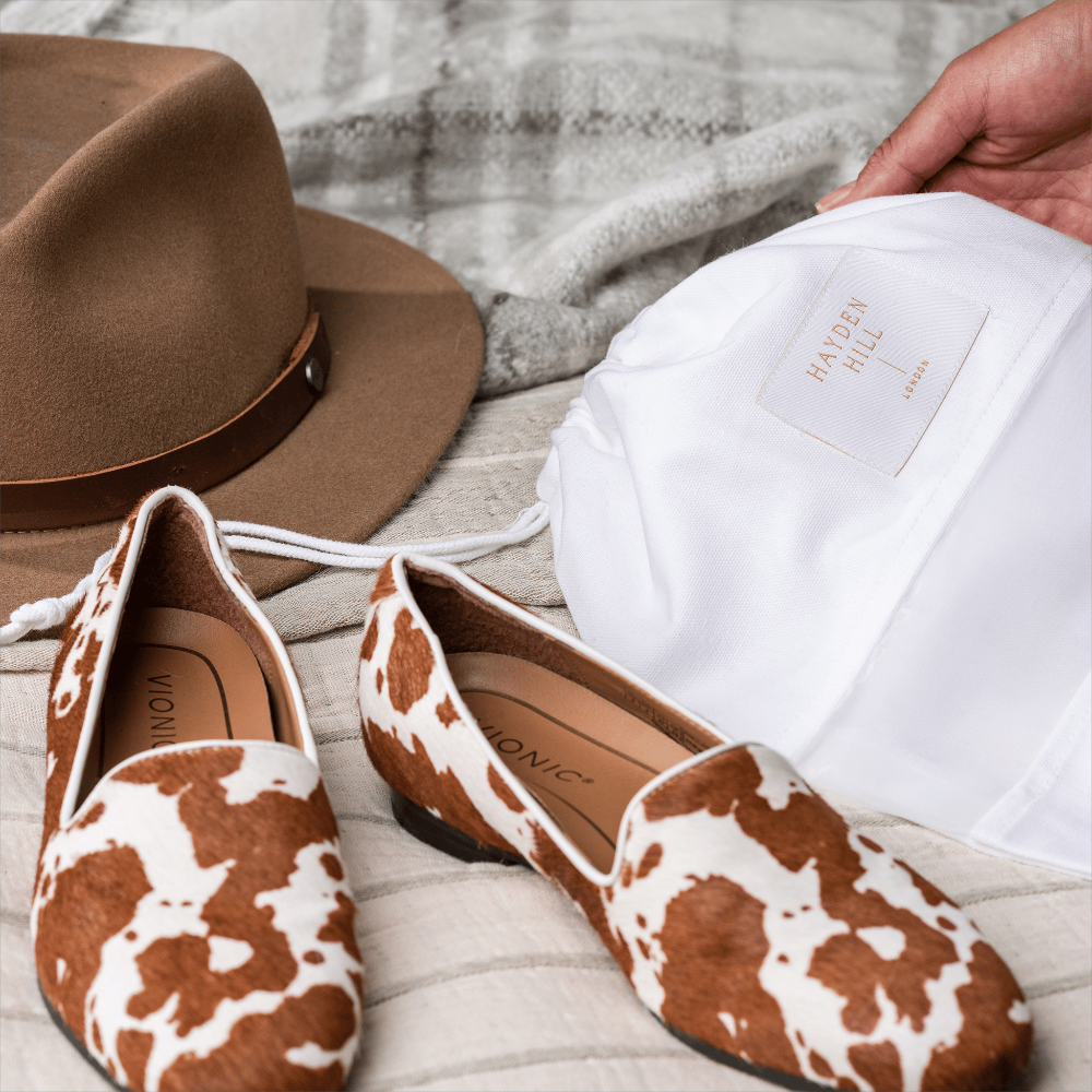 A pair of women's slip-on flats with a brown and white cow-print pattern, displayed next to a white dust bag labeled 'HAYDEN HILL' and a brown felt hat on a neutral fabric background.