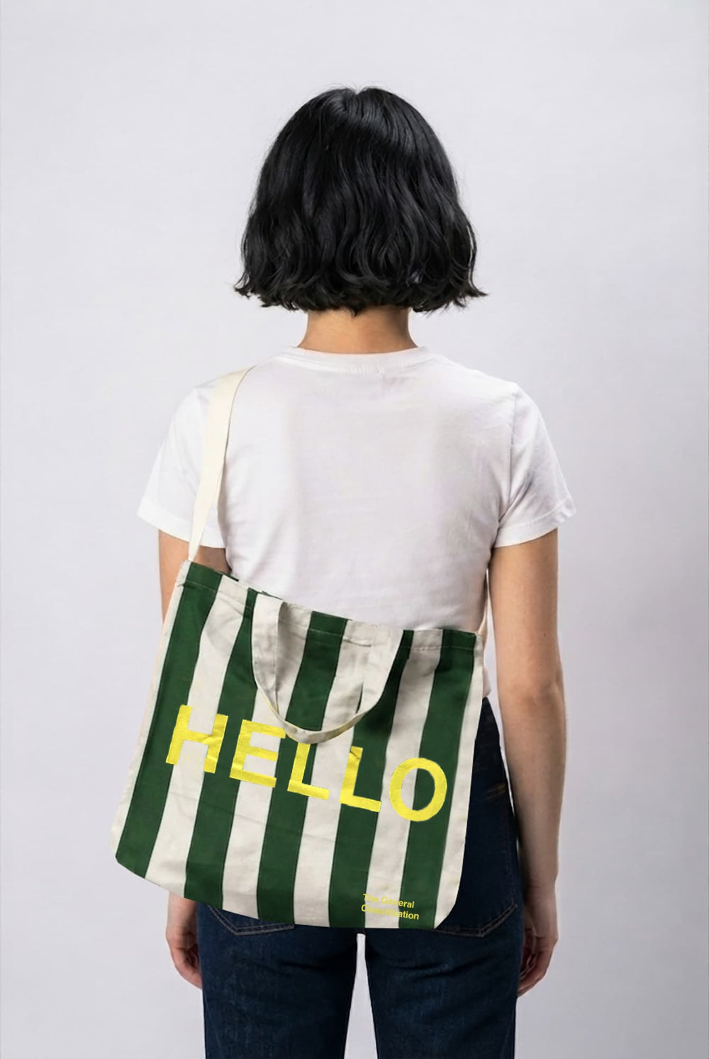 A person shown from behind wearing a white t-shirt and dark jeans, carrying a green and white striped canvas musette bag with yellow 'HELLO' text on the back.
