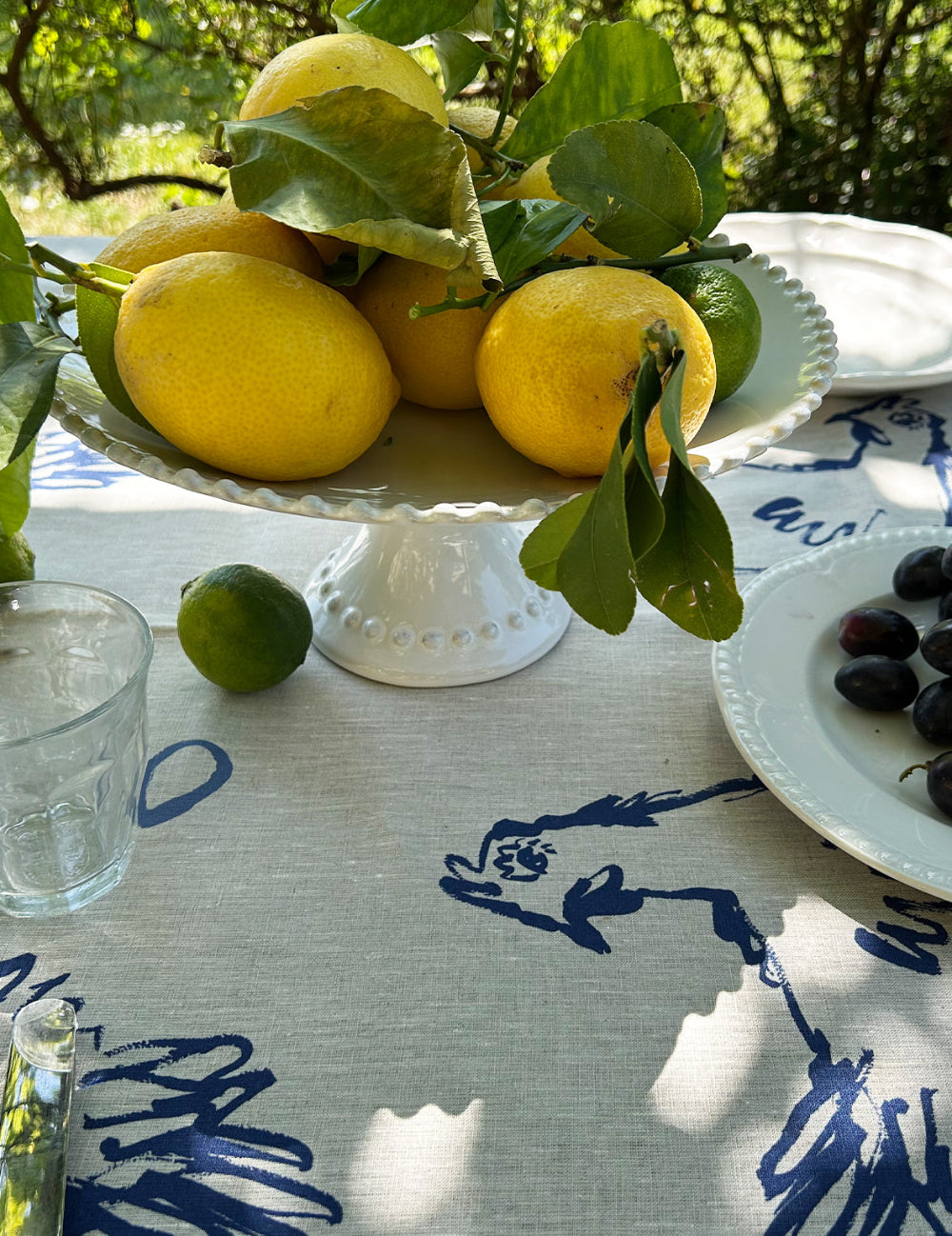 Lemons on a white pedestal dish with a blue and white linen tablecloth in the background