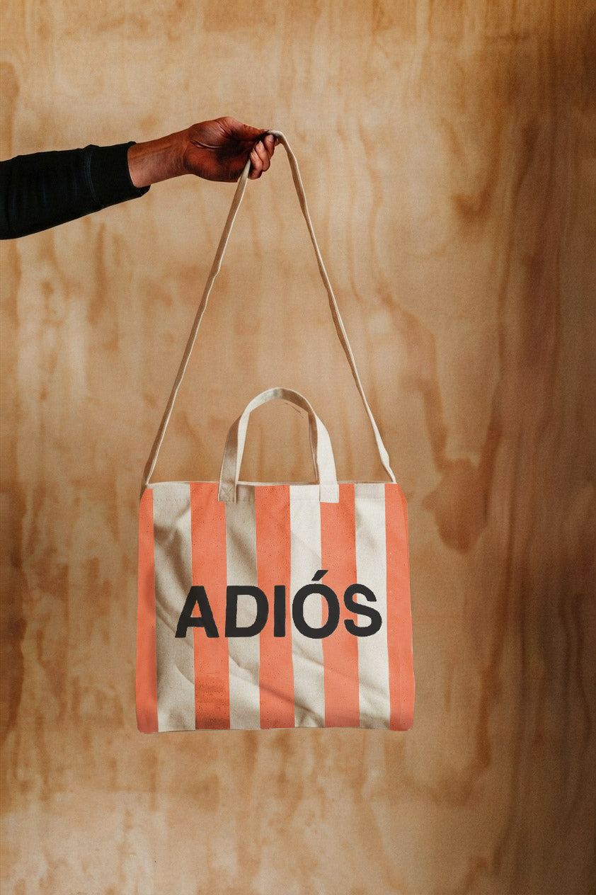 A striped canvas tote bag with coral pink and white vertical stripes, featuring the word 'ADIÓS' printed in black across the front. A hand holding a black sleeve is holding the top strap of the bag against a light wood panel background.