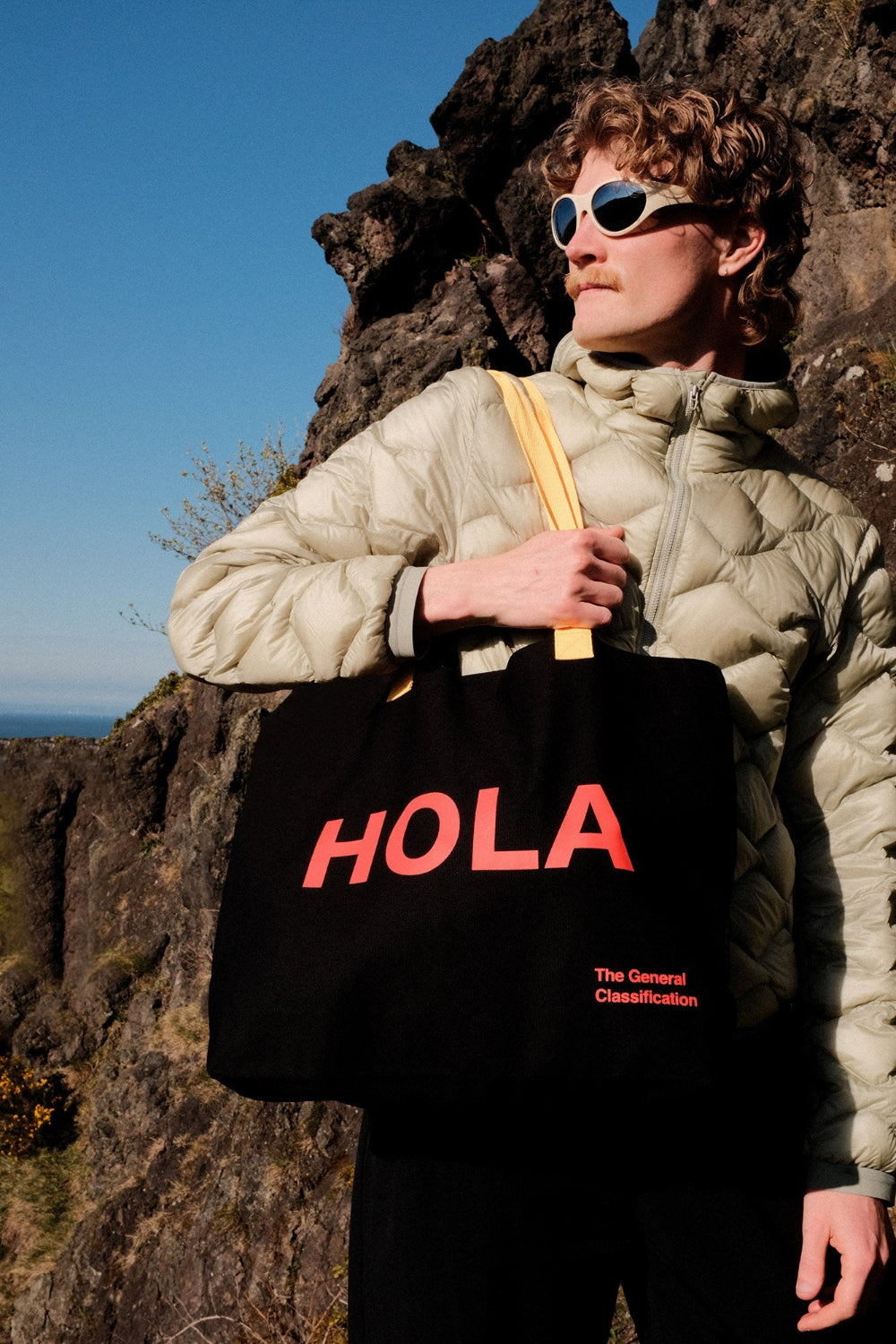 Person holding a black tote bag with 'HOLA' in red text, standing on rocky terrain.