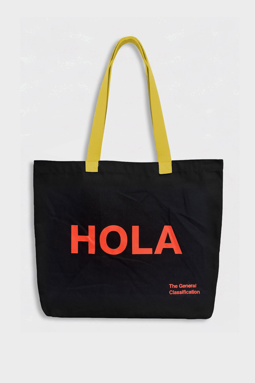 A black canvas tote bag with bright yellow shoulder straps and bold orange-red 'HOLA' text on the front, with smaller orange-red text reading 'The General Classification' below, shown against a light gray background.